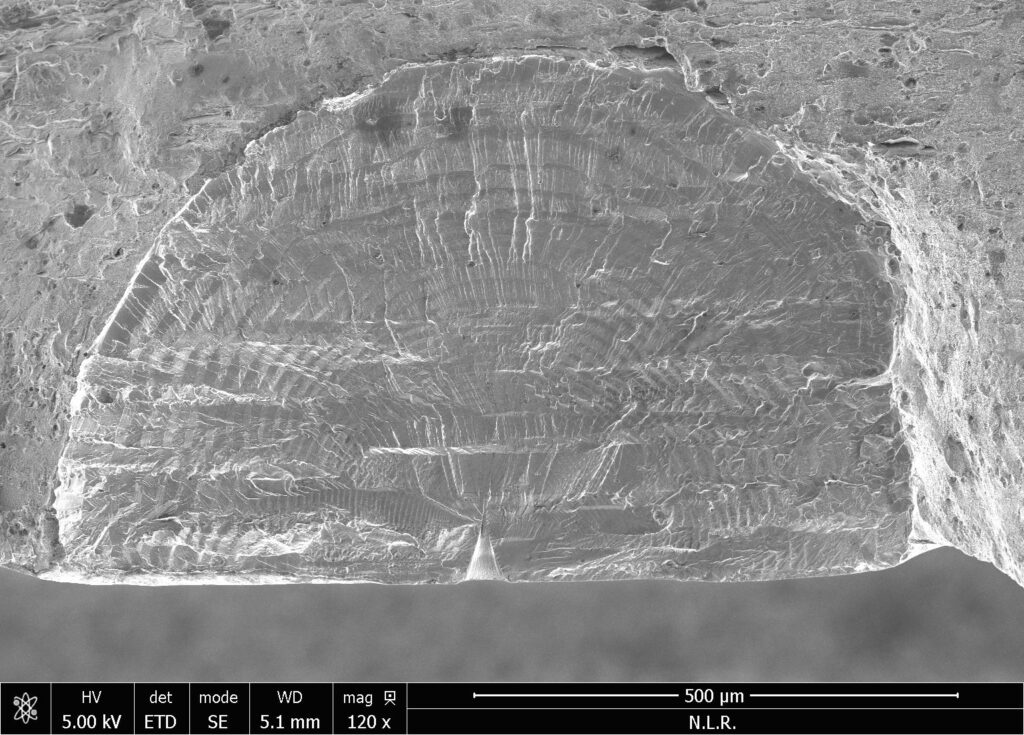 Foto van een vermoeiingsbreukvlak gemaakt met een elektronenmicroscoop (120x vergroot). De vermoeiingsbreuk is gegroeid vanuit een kunstmatig defect dat met een laser is aangebracht op het oppervlak van het proefstuk.